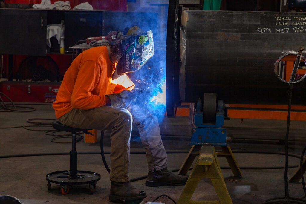 A welder wearing an orange shirt and a welding mask works on a weld.