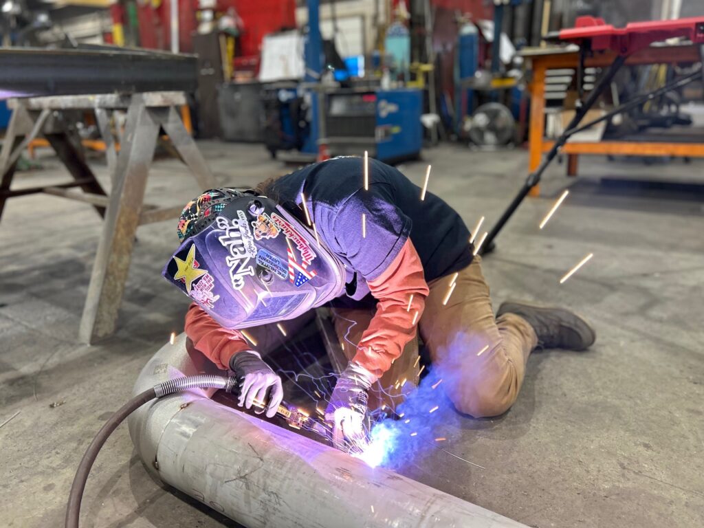 A welder using a MIG welding gun joins metal components in the GSM Industrial fabrication shop, surrounded by bright sparks and protective gear.