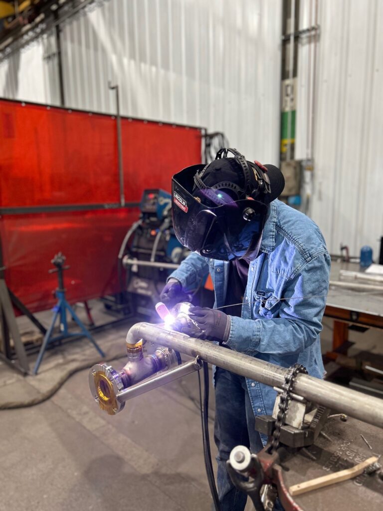 A welder performs TIG welding on stainless steel piping in GSM Industrial’s fabrication shop, using a torch and filler rod for precise, high-quality joints.